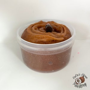 May include: A clear plastic container filled with brown slime with a swirl pattern and a small black bead on top.
