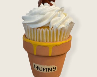 May include: A white cupcake with chocolate frosting and a chocolate topping, sitting in a small terracotta pot with yellow paint and the word "Hunny" written on it.