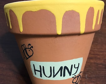May include: A terracotta flower pot with a yellow glaze dripping down the sides. The pot is decorated with a black bee and the word "Hunny" written in black.
