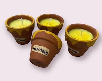 May include: Four small terracotta flower pots with yellow wax candles inside. The wax is dripping down the sides of the pots. The pots are labeled "Hunny".