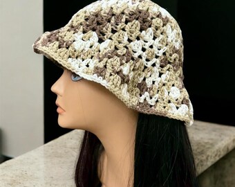 May include: A crocheted bucket hat in a neutral color palette of white, beige, and brown. The hat features a wide brim and a textured, open weave design. The hat is displayed on a mannequin head.