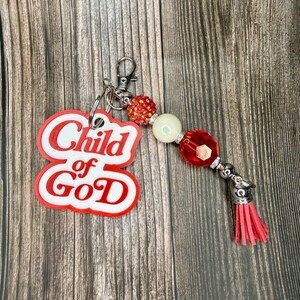 May include: A keyring featuring a white and red acrylic charm with the text Child of God in a bold font. The charm is attached to a silver metal ring with a series of beads including a red rhinestone bead, a cream bead, and a faceted red bead, ending with a pink faux leather tassel.