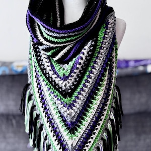 May include: A crocheted triangular shawl with a black, white, green, and purple color scheme. The shawl has a fringe border and is displayed on a white mannequin.