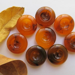 May include: Nine round buttons in shades of amber and brown, each with four holes. The buttons have a marbled effect. A dried, yellow leaf lies near the buttons on a white surface. These buttons are suitable for sewing and craft projects.