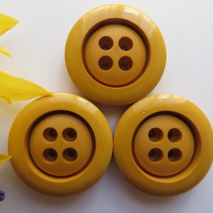 May include: Three round, mustard-yellow buttons with four holes each. The buttons are arranged on a white surface, with yellow flower petals and purple flower petals in the background.