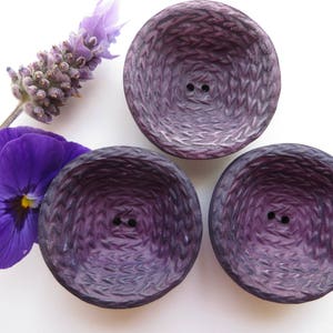 Peut inclure: Trois boutons en tissu violet avec une texture tissée et deux trous chacun. Les boutons sont disposés en forme de triangle sur un fond blanc.