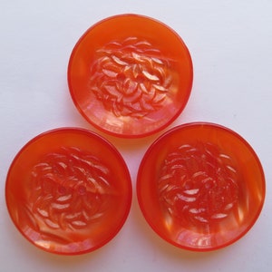 May include: Three round, orange buttons with a floral design in the center. The buttons are made of a translucent material, and the floral design is raised. The buttons are arranged on a white surface.