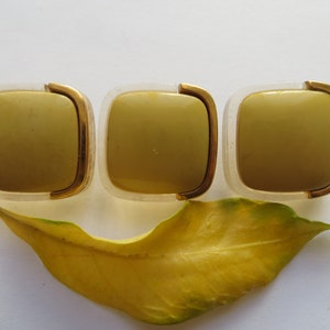 May include: Three square-shaped, gold-rimmed, clear plastic containers with a light green interior. The containers are arranged in a row.