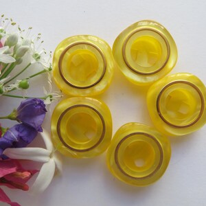 May include: Six yellow plastic buttons with a gold trim around the edge. The buttons have a circular hole in the center.
