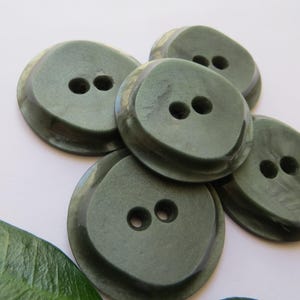 Puede incluir: Primer plano de varios botones verde oliva con dos agujeros cada uno. Los botones tienen una forma redondeada ligeramente irregular y un acabado brillante. Una hoja verde es visible en la esquina inferior izquierda. Estos botones son adecuados para proyectos de costura.