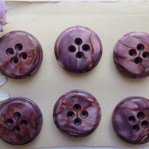 Peut inclure: Six boutons ronds marbrés dans des tons de violet et de bordeaux, chacun avec quatre trous. Les boutons sont disposés sur une surface crème, avec une branche de lavande et une marguerite en arrière-plan.
