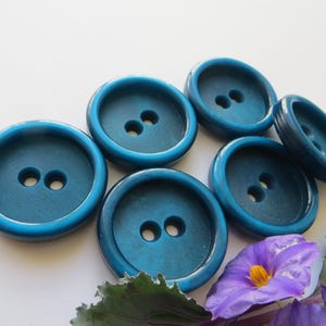 Op de afbeelding: Zes tealblauwe plastic knoppen met elk twee gaten. De knoppen zijn rond en hebben een glanzende afwerking.