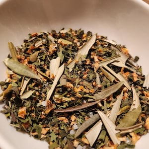 Puede incluir: Un bol blanco lleno de una mezcla de hierbas secas, que incluyen hojas verdes, cáscara de naranja y hojas de olivo.