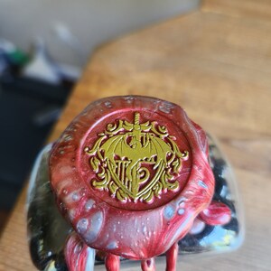 May include: A red and silver wax seal with a gold crest featuring a winged creature and a sword. The crest is surrounded by a decorative border.