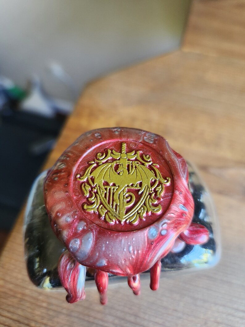 May include: A red and silver wax seal with a gold crest featuring a winged creature and a sword. The crest is surrounded by a decorative border.