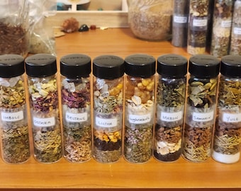May include: Eight small glass jars with black lids, each filled with different dried herbs and labeled with the names of eight different holidays: Imbolc, Ostara, Beltane, Litha, Lughnasadh, Mabon, Samhain, and Yule.