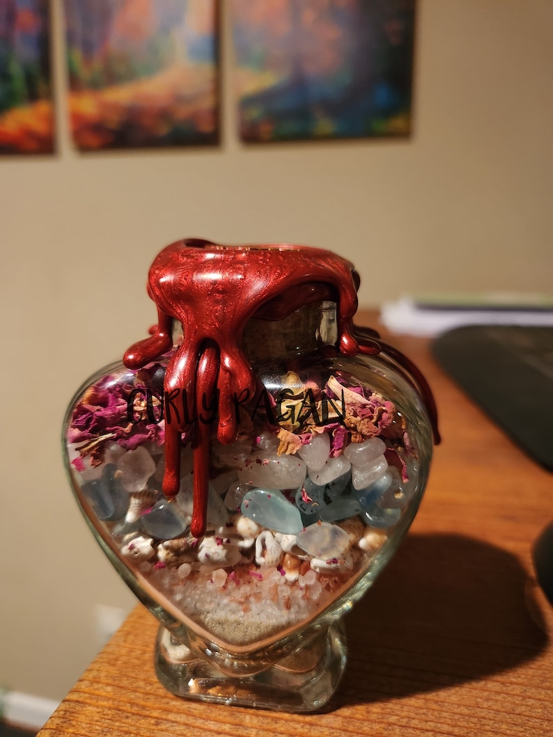 Op de afbeelding: Een hartvormige glazen fles gevuld met roze en witte rozenblaadjes, heldere en blauwe kristallen en wit zand. De fles is verzegeld met een rode waszegel die langs de zijkant druppelt. De fles is gelabeld met "Curly Pagan".
