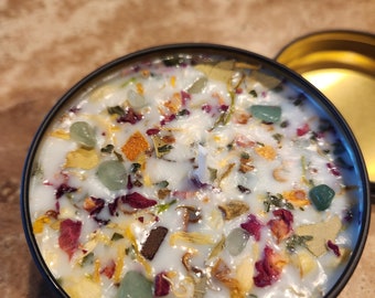 May include: A white candle with a variety of dried flowers, herbs, and green stones embedded in the wax. The candle is in a black metal container.
