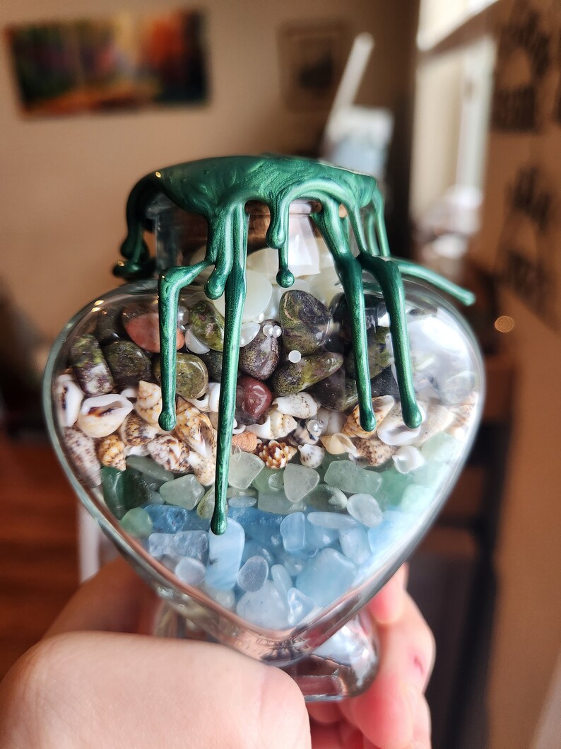 May include: A heart-shaped glass jar filled with various small stones and seashells. The jar is sealed with a green wax seal that drips down the sides.