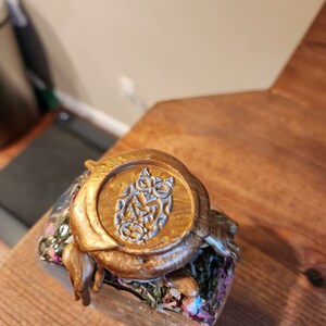 May include: A glass jar filled with dried herbs and flowers, sealed with a gold wax seal featuring a silver owl design.