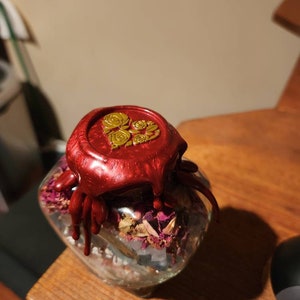 Op de afbeelding: Een glazen pot gevuld met gedroogde rozenblaadjes en andere kruiden, verzegeld met een rode waszegel met een gouden bloemenmotief.