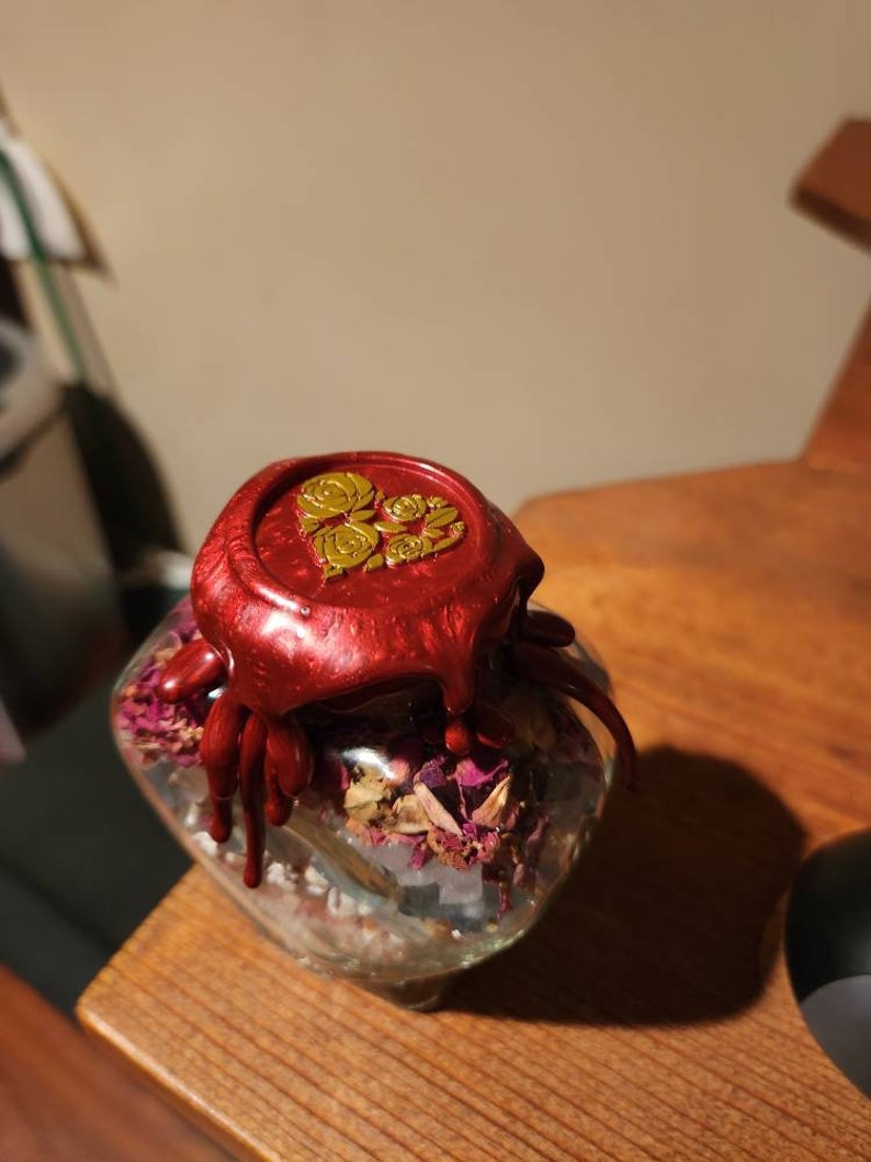 Op de afbeelding: Een glazen pot gevuld met gedroogde rozenblaadjes en andere kruiden, verzegeld met een rode waszegel met een gouden bloemenmotief.