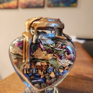 May include: A heart-shaped glass jar filled with various herbs, crystals, and gold leaf. The jar is sealed with a gold wax seal and has the text "Curly Pagan" on the front.