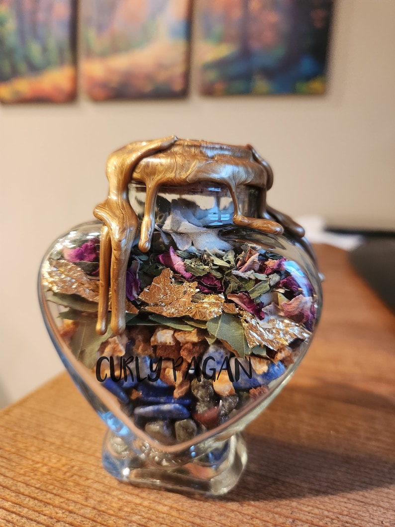 May include: A heart-shaped glass jar filled with various herbs, crystals, and gold leaf. The jar is sealed with a gold wax seal and has the text "Curly Pagan" on the front.