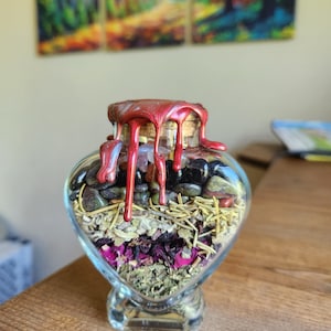 May include: A heart-shaped glass bottle filled with herbs, crystals, and dried flowers. The bottle is sealed with a red wax seal that is dripping down the sides.