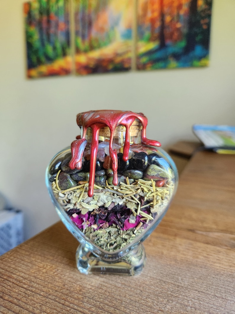 May include: A heart-shaped glass bottle filled with herbs, crystals, and dried flowers. The bottle is sealed with a red wax seal that is dripping down the sides.