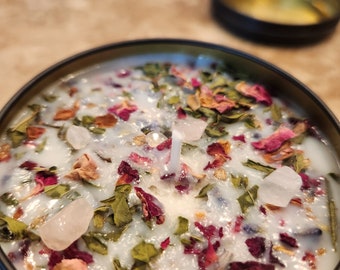 May include: A black candle tin filled with a light blue wax, adorned with dried rose petals, green herbs, and clear quartz crystals. A white wick is centered in the wax. The lid is off, revealing the candle's contents.