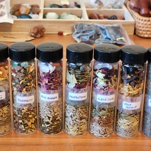 Op de afbeelding: Acht kleine glazen flesjes met zwarte deksels gevuld met verschillende kruiden, kristallen en andere ingrediënten. Elk flesje heeft een etiket met een andere naam, waaronder "Empowerment", "Prosperity", "Rid Anger", "Protection", "Self-Love", "Stop Gossip" en "All-Purpose Incense".
