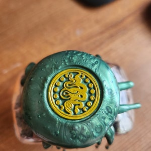 May include: A green bottle cap with a gold snake design. The cap has a textured surface and a shiny finish.