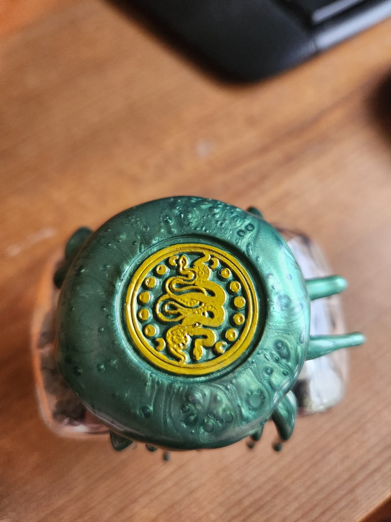 May include: A green bottle cap with a gold snake design. The cap has a textured surface and a shiny finish.