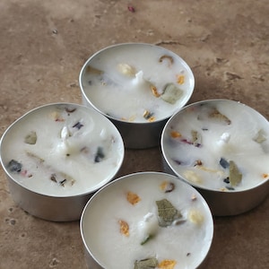 May include: Four white tea light candles with dried flowers and herbs embedded in the wax. The candles are in silver holders.