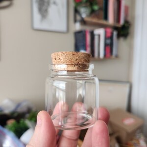 May include: A small, clear glass bottle with a cork stopper. The bottle is empty and has a rounded bottom.