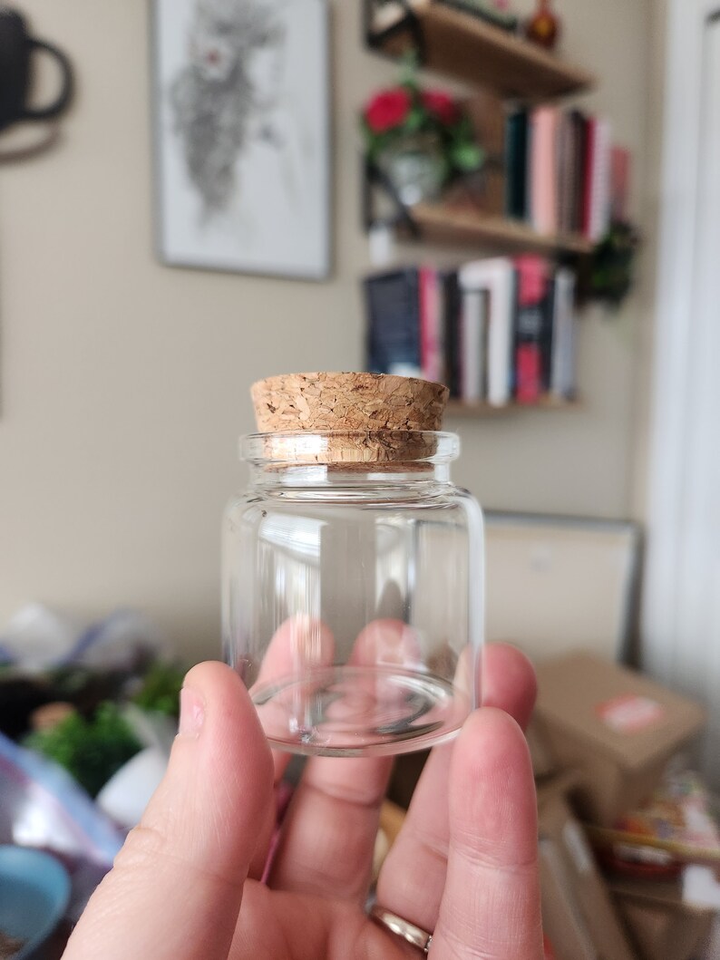 May include: A small, clear glass bottle with a cork stopper. The bottle is empty and has a rounded bottom.