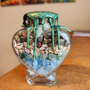 May include: A heart-shaped glass jar filled with various stones, shells, and beads. The jar is sealed with a green wax lid that has drips running down the sides.