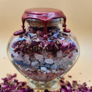 Op de afbeelding: Een hartvormige glazen fles gevuld met roze en blauwe kristallen, rozenblaadjes en een rode waszegel. De fles is gelabeld met "GORKY PAGAN".