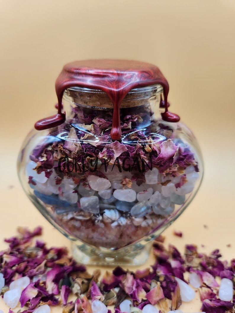 Op de afbeelding: Een hartvormige glazen fles gevuld met roze en blauwe kristallen, rozenblaadjes en een rode waszegel. De fles is gelabeld met "GORKY PAGAN".