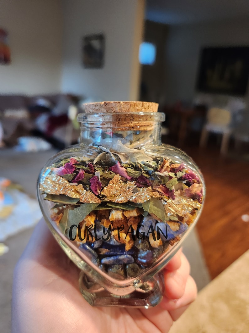 May include: A heart-shaped glass bottle filled with dried herbs, flowers, and gold leaf. The bottle has a cork stopper and a label that reads "Curly Pagan".