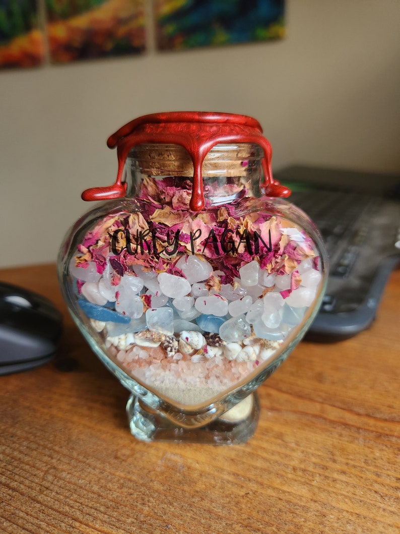 Op de afbeelding: Een hartvormige glazen pot met een rode waszegel. De pot is gevuld met roze rozenblaadjes, witte kristallen, blauwe stenen en wit zand. De pot heeft een etiket met de tekst "Curly Pagan".