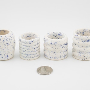 Może przedstawiać: Cztery małe, białe ceramiczne pojemniki z niebieskimi plamkami. Pojemniki mają różne kształty, w tym cylindryczny i żebrowany. Moneta jest umieszczona przed pojemnikami w celu porównania rozmiaru.