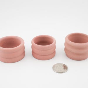 May include: Three small, pink, ceramic pots with a ridged design. The pots are arranged in a row, varying slightly in size. A coin is placed in front of the pots for scale. The pots are likely for small plants or decorative use.