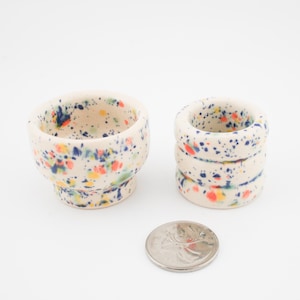 Peut inclure: Deux pièces en céramique avec un motif de peinture mouchetée bleue, rouge et jaune. L'une est un petit bol, l'autre est une pile de trois anneaux. Une pièce de monnaie est à côté des pièces pour la comparaison de taille.