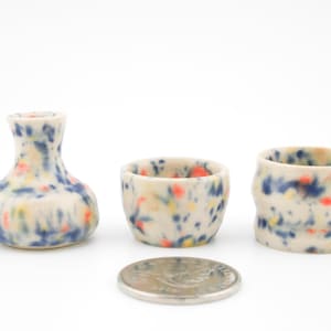 May include: A set of three small ceramic items with a speckled pattern. The set includes a small vase and two rounded cups. The pieces are white with blue, orange, and yellow speckles. A coin is in the foreground for size comparison.