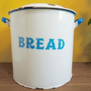 May include: White enamel bread bin with a lid and blue detailing. The word "BREAD" is written in blue, vintage-style lettering on the front. The bin has blue handles and a blue rim around the lid.
