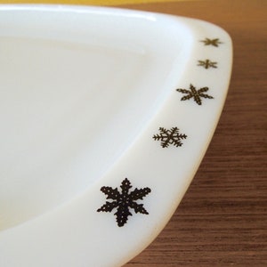 Op de afbeelding: Witte keramische schaal met een sneeuwvlokkenontwerp rond de rand. De sneeuwvlokken zijn zwart en variëren in grootte.
