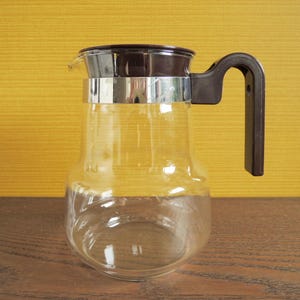 Peut inclure: Une carafe à café en verre transparent avec un couvercle et une poignée en plastique marron, et une bande métallique argentée. La carafe est posée sur une surface en bois avec un fond jaune.