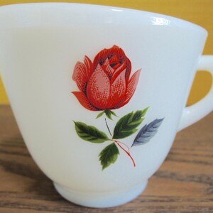 Può includere: Lattiera color crema con un motivo a rosa rossa. La rosa ha petali rossi e foglie verdi. La lattiera ha un manico e un beccuccio. Un articolo per la cucina in stile vintage.